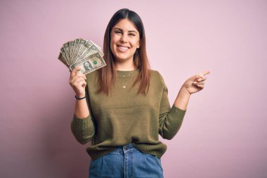 Soyutlanmış pembe arka planda dolarlar tutan genç ve güzel bir kadın. Elleri ve parmakları kenarda mutlu bir şekilde işaret ediyor.
