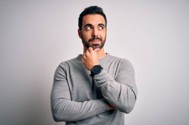 Sakallı, sıradan bir süveter giyen, eli çenesinde beyaz arka planda soru soran, düşünceli bir genç adam. Düşünceli bir yüzle gülümsüyor. Şüphe kavramı.