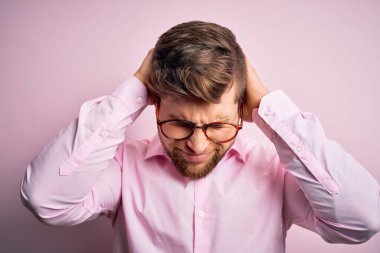 Sakallı, mavi gözlü, pembe gömlekli, gözlüklü, baş ağrısı çeken, çaresiz ve stresli genç sarışın bir adam. Ağrı ve migren yüzünden. Ellerini başının üstüne koy..