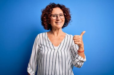 Orta yaşlı, kıvırcık saçlı, rahat çizgili tişört giyen mutlu yüzüyle gülümseyen ve baş parmağıyla yüzünü gösteren bir kadın..