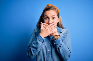 Mavi arka plan üzerine günlük kot gömlek ve diyabet giyen genç, güzel sarışın kadın yanlışlıkla ağızlarını kapatarak şok oldu. Gizli konsept.