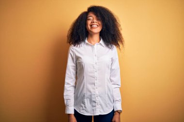 Afro saçlı, güzel, Afro-Amerikan zarif genç bir kadın yüzünde mutlu ve havalı bir gülümsemeyle sarı arka planda duruyor. Şanslı kişi..