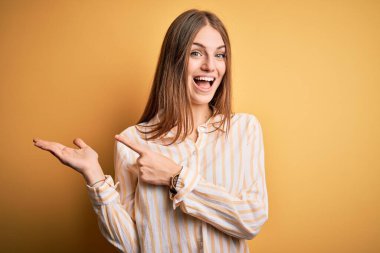 İzole edilmiş sarı arka plan üzerinde sıradan çizgili tişört giyen genç ve güzel bir kızıl kadın kameraya hayret ediyor ve gülümsüyor..