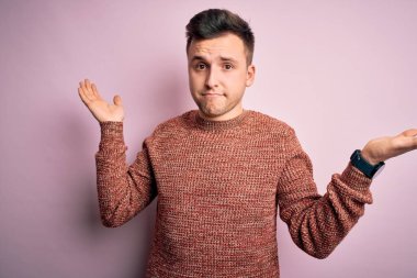 Pembe izole arka plan üzerine sıradan bir kazak giyen yakışıklı beyaz bir adam. Kolları ve elleri havada, kafası karışmış bir yüz ifadesi. Şüphe kavramı.