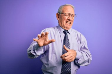 Gri saçlı, gözlüklü kıdemli iş adamı mor izole edilmiş arka planda duruyor. İğrenç bir ifade, tiksinti reaksiyonu yüzünden tiksinti ve korku ifadesi takınıyor. Eller havada