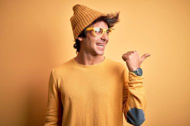 Genç ve yakışıklı bir adam, günlük tişört ve gözlük takıyor, sarı arka planda, mutlu yüzüyle gülümsüyor ve başparmağıyla yan tarafı işaret ediyor..