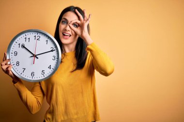 Mavi gözlü genç bir kadın geri sayım yapıyor. Sarı arka planda büyük bir saat tutuyor. Gülümsüyor.