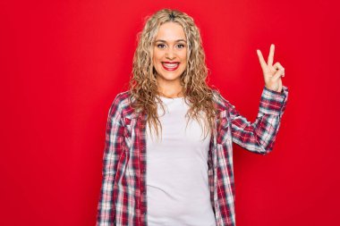 İzole edilmiş kırmızı arka planda dikilmiş, sıradan bir tişört giyen genç, sarışın bir kadın zafer işareti yaparken parmaklarını kameraya doğru gülümsüyor. İki numara..