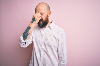 Soyutlanmış pembe arka plan üzerinde zarif bir gömlek giyen sakallı yakışıklı kel adam yorgun burnu ve gözleri ovuşturup yorgun ve baş ağrısı çekiyor. Stres ve hüsran kavramı.