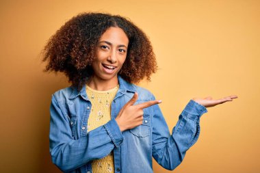 Afro saçlı, sarı arka planda sıradan bir kot gömlek giyen genç Afro Amerikalı kadın kameraya hayret ediyor ve gülümsüyordu..