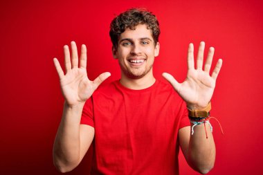 Sarışın, kıvırcık saçlı, kırmızı arka planda sıradan tişörtler giyen genç ve yakışıklı bir adam on numarayı gösterirken kendinden emin ve mutlu bir şekilde gülümsüyor..