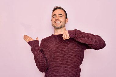 Mavi gözlü, sıradan bir süveter giyen genç bir adam pembe arka planda durup hayrete düşmüş ve kameraya gülümserken eliyle ve parmağıyla işaret ediyor..