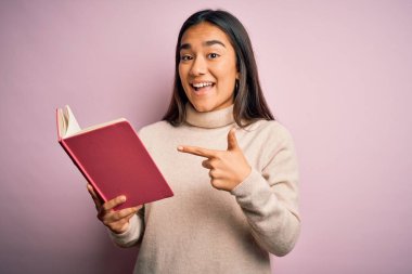 Genç Asyalı akıllı kadın kitap günlüğü tutuyor pembe arka planda duruyor. El ve parmağıyla işaret ederek çok mutlu.