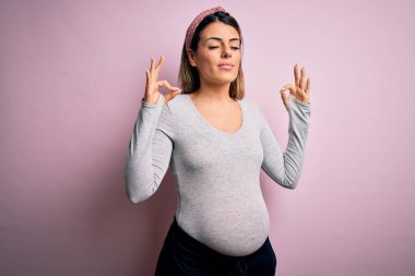 Genç, güzel, esmer bir kadın hamile, pembe arka planda bebek bekliyor. Rahat ol ve gözün kapalı gülümse. Parmaklarınla meditasyon hareketi yap. Yoga konsepti.