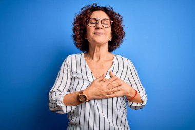 Orta yaşlı güzel kıvırcık saçlı kadın izole edilmiş arka planda sıradan çizgili tişört giyiyor. Elleri göğsünde, gözleri kapalı ve yüzünde minnet dolu bir jest var. Sağlık kavramı.