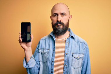 Sakallı yakışıklı kel adam. Akıllı telefon tutuyor. Sarı arka planda ekranı gösteriyor.