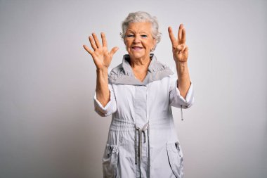 Son sınıf güzeli, gri saçlı, sıradan bir ceket giyen, beyaz arka plan üzerinde duran ve 8 numarayı gösteren, kendinden emin ve mutlu gülümseyen bir kadın..