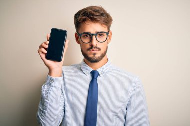 Akıllı telefonu elinde tutan genç iş adamı, izole edilmiş beyaz arka planda duruyor. Akıllı bir yüz ifadesiyle ciddi olduğunu düşünüyor.