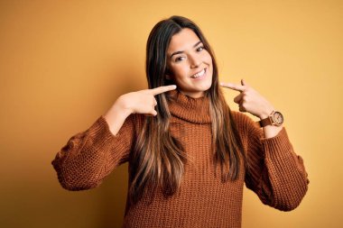 İzole edilmiş sarı arka planda dikilen sıradan bir süveter giyen neşeli bir şekilde gülümseyen ve parmaklarıyla ve ağzıyla işaret eden genç bir kız. Diş sağlığı konsepti.