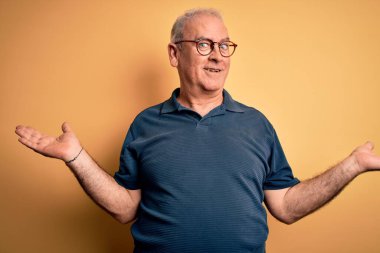 Orta yaşlı, yakışıklı, polo giyen ve sarı arka planda gözlük takan yakışıklı adam gülümseyerek avuçlarını açıyor, sunuyor, reklam yapıyor, karşılaştırıyor ve dengede duruyor.