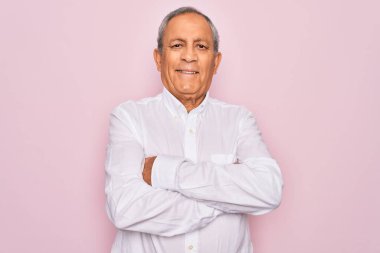 Soyutlanmış pembe arka planda zarif gömlekler giyen yaşlı, gri saçlı, kollarını kavuşturup kameraya bakan yakışıklı bir adam. Pozitif kişi.