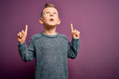 Mor arka plan üzerinde duran mavi gözlü küçük beyaz çocuk şaşırmış ve şaşırmış bir şekilde yukarı bakıyor ve parmaklarıyla işaret ediyor ve kollarını kaldırıyordu..