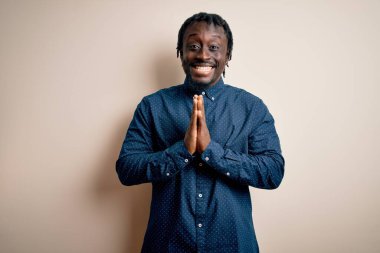 Genç, yakışıklı, Afro-Amerikan bir adam, günlük gömleğini giymiş, beyaz arka planda durmuş, ellerini birleştirip af diliyor, kendinden emin bir şekilde gülümsüyor..
