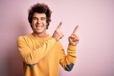 Sarı günlük tişört giyen yakışıklı genç adam izole edilmiş pembe arka planda duruyor gülümsüyor ve kameraya bakıyor iki eliyle ve parmaklarıyla yan tarafı işaret ediyor..
