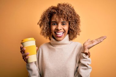 Genç Afro-Amerikan kıvırcık kadın sarı arka planda bir fincan kahve içiyor mutlu ve heyecanlı, kazanan ifade zaferi kutluyor büyük bir gülümsemeyle ve el kaldırarak.