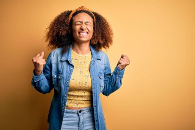 Güzel Afro saçlı Afrikalı Amerikalı, sarı izole edilmiş arka planda mutlu ve heyecanlı bir şekilde kollarını kaldırıp, gülümseyerek ve başarı çığlıkları atarak zafer jesti yapıyor. Kutlama konsepti.