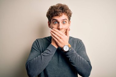 Sarışın, kıvırcık saçlı, beyaz arka planda günlük süveter giyen, ağzı yanlışlıkla ellerle kapatan genç, yakışıklı bir adam. Gizli konsept.