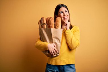 Genç ve güzel bir kadın sarı arka planda bir torba taze sağlıklı ekmek tutuyor. Elleri çenesinde soru sorarken, düşünceli bir ifade takınıyor. Gülen ve düşünceli bir yüz. Şüphe kavramı.