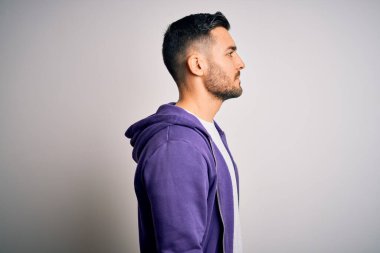 Mor eşofman üstü giyen genç yakışıklı adam, izole edilmiş beyaz arka planda duruyor, yan tarafa bakıyor, rahat bir görünüm sergiliyor, kendine güvenen bir gülümsemeyle doğal bir yüz ifadesi var..
