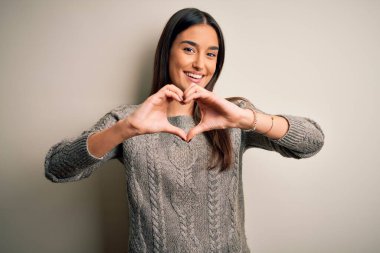 Genç, güzel esmer kadın, izole edilmiş beyaz arka plan üzerine gündelik süveter giyip, elleriyle kalp sembolü yapan, aşkla gülümsüyor. Romantik konsept.