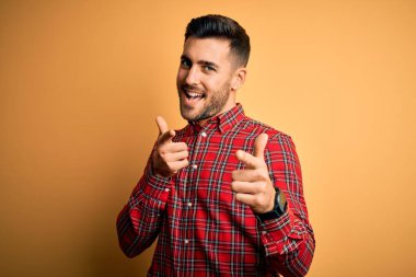 Genç yakışıklı adam rahat gömlek giyen izole sarı arka plan üzerinde duran mutlu ve komik yüzü ile kameraya parmakları işaret. İyi enerji ve titreşimler.