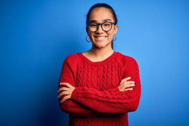 Genç, güzel Asyalı kadın mavi arka planda gözlük takıyor ve kollarını kavuşturup kameraya gülümsüyor. Pozitif kişi.