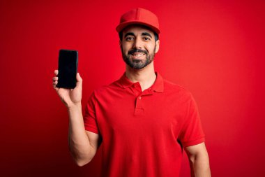 Sakallı yakışıklı teslimatçı elinde akıllı bir telefonla gülen bir gülümsemeyle ayakta duran ve dişlerini gösteren bir yemek uygulaması sunuyor.