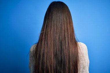 Genç ve güzel Asyalı kadın, günlük süveter giyip mavi izole arka planda dikiliyor ve kollarını kavuşturup arkasını çeviriyor.