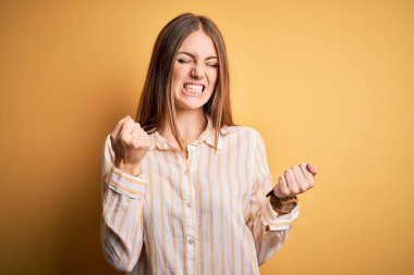 İzole edilmiş sarı arka plan üzerine sıradan çizgili tişört giyen genç ve güzel bir kızıl kadın. Kolları havada ve gözleri kapalı zaferi kutlarken gülümsüyor. Kazanan konsept.