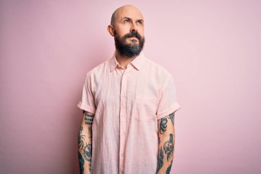 Sakallı ve dövmeli yakışıklı, pembe arka planda günlük gömleği olan, yüzünde gülümseme ve doğal ifadeyle yan yana bakan kel bir adam. Kendinden emin gülen.