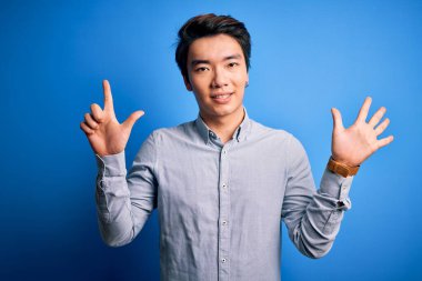 Genç yakışıklı Çinli bir adam, günlük gömleğini giymiş, mavi arka planda dikiliyor ve yedinci parmaklarını gösterirken, kendinden emin ve mutlu bir şekilde gülümsüyor..