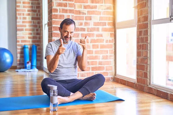 Middle age handsome sportman sitting on mat doing stretching yoga exercise at gym smiling doing talking on the telephone gesture and pointing to you. Call me.