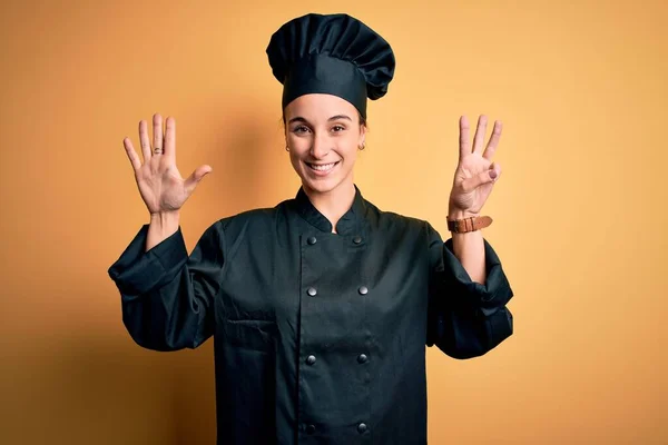 年轻美丽的厨师女士身穿炊事服 头戴一顶帽子 站在黄色的背景上 露出八号手指 面带微笑 自信而快乐 — 图库照片