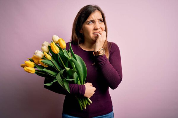 Young blonde woman holding romantic bouquet of yellow tulips flowers over pink background looking stressed and nervous with hands on mouth biting nails. Anxiety problem.