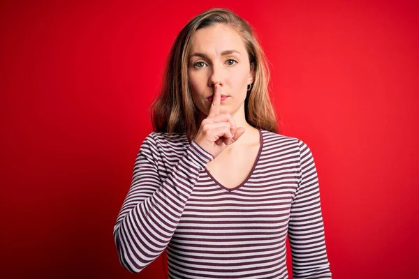唇に指で静かにして欲しいと願う孤立した赤い背景にカジュアルなストライプのTシャツを着た若い美しいブロンドの女性 沈黙と秘密の概念 — ストック写真