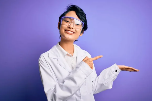 紫の背景にコートと眼鏡を身に着けている若い美しいアジアの科学者の女の子は驚いて 手で提示しながら カメラに笑顔と指で指して — ストック写真