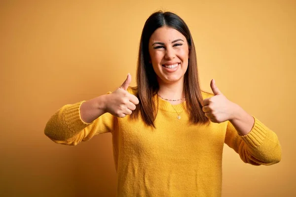黄色の隔離された背景の成功の印の上にカジュアルなセーターを身に着けている若い美しい女性は手で積極的なジェスチャーを行う 親指を笑顔と幸せ 陽気な表情と勝者のジェスチャー — ストック写真
