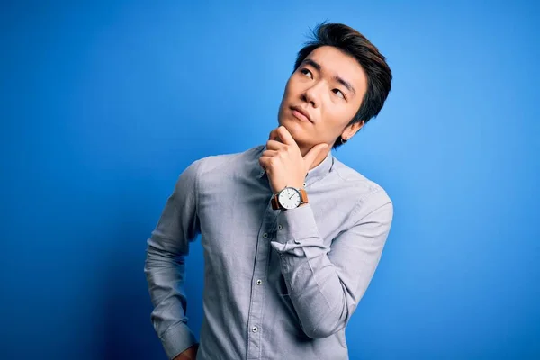 Young handsome chinese man wearing casual shirt standing over isolated blue background with hand on chin thinking about question, pensive expression. Smiling with thoughtful face. Doubt concept.