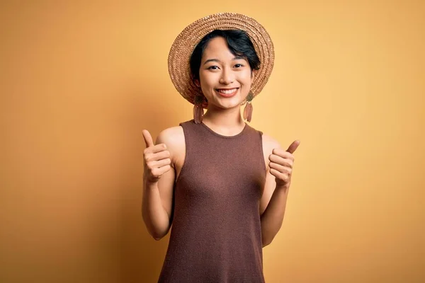 年轻美丽的亚洲女孩穿着休闲装 头戴帽子 戴黄色背景的孤立的成功标志 手拿着积极的手势 竖起大拇指 笑容满面 快乐的表情和胜利的姿态 — 图库照片
