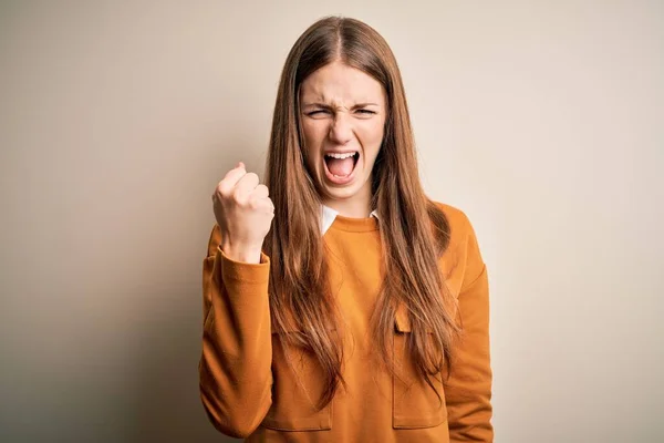 Young beautiful redhead woman wearing casual sweater over isolated white background angry and mad raising fist frustrated and furious while shouting with anger. Rage and aggressive concept.